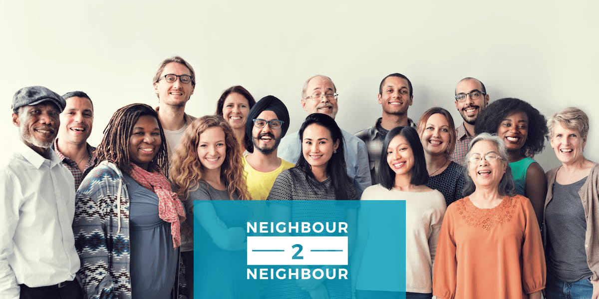 a photo of a group of diverse people are standing closely, looking and smiling at the camera. the neighbour to neighbour logo is seen in the image.